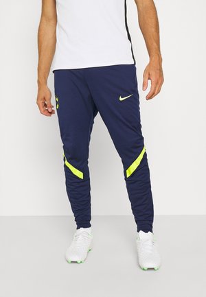 Mann trägt marineblaue Nike-Sporthose mit gelben Akzenten und weißen Fußballschuhen, steht vor einem schlichten weißen Hintergrund.