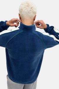 Pullover in pile blu navy con colletto alto, caratterizzato da strisce laterali bianche e dettagli verdi sulle cuciture e sui polsini. Tessuto morbido e testurizzato.