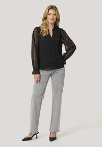 Svart spetsblus med långa ärmar, V-ringning och geometriska mönster. Matchas med ljusgrå raka jeans och svarta klacktofflor.