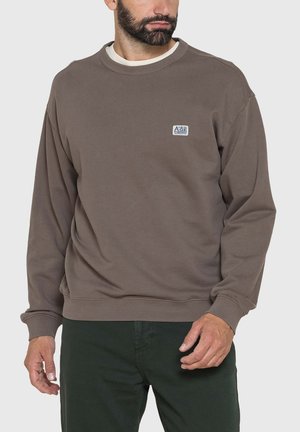 Uomo che indossa una felpa marrone larga con scollo a girocollo e un piccolo logo rettangolare sul petto, e pantaloni verde scuro, in piedi davanti a uno sfondo semplice.