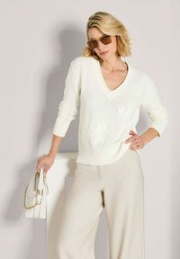 Vrouw in een crèmekleurige trui en wijde broek, met een witte handtas en oversized bruine zonnebril, staand tegen een lichte achtergrond.