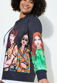 Námořnická mikina s kapsou na zip a třemi ilustrovanými ženami na rukávech, které mají květinové, pruhované a vzorované designy v živých barvách.