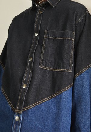 Chemise en denim avec boutons-pression, partie supérieure noire, partie inférieure bleue, poche poitrine et coutures contrastées visibles.