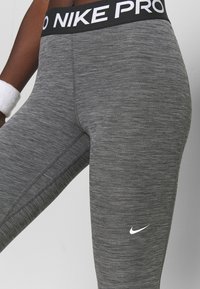 Zbliżenie osoby noszącej szare legginsy Nike Pro z białym logo Swoosh i czarnym pasem w talii, noszącej białą opaskę na nadgarstek.