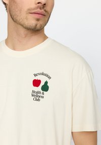 Crème kortom shirt met rode appel- en groene peerafbeeldingen en tekst "Revolution Health & Wellness Club" in zwart. Katoenen materiaal.