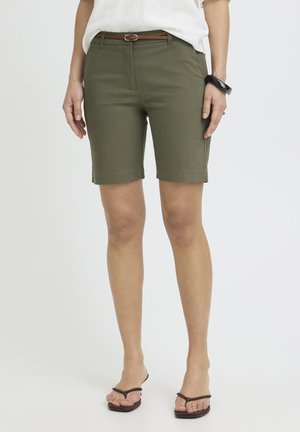 Persoon draagt olijfgroene knielange shorts met een smalle bruine riem, een wit overhemd in de broek gestopt, een zwarte armband en bruine teenslippers.