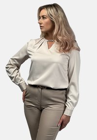 Kremowa bluzka z dekoltem w kształcie łezki, długimi rękawami i plisowanymi detalami. W zestawie z jasnobeżowymi spodniami i czarnym paskiem.