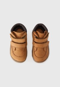 Kleine tan kleurige peuterschoenen met twee klittenbandsluitingen en donkere, gewatteerde kraag, gezien van bovenaf op een witte achtergrond.