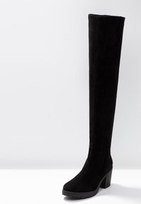 Botas negras de gamuza por encima de la rodilla, con un Tacón ancho y suela de goma, que presentan un diseño elegante y aerodinámico sin herrajes visibles.