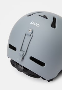 Pelēks ķivere ar ventilācijas atverēm, regulējamu siksnu un POC logotipu; matēta apdare ar teksturētu virsmu un aizmugurē esošu sprieguma detaļu.