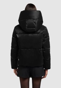 Črna puhovka z veliko kapuco, prešito zasnovo, sijajnim zaključkom in izrazitimi vodoravnimi šivi. Kratka dolžina, kombinirana s črnimi kratkimi hlačami.