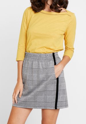 Vrouw draagt een geel shirt met lange mouwen en een hooggetailleerde zwart-witte geruite rok met een zwarte streep aan de zijkant, hand in de zak van de rok.