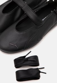 Chaussures en cuir noir style Mary Jane avec logo Puma et deux paires de lacets en ruban satiné noir sur fond blanc.