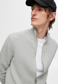 Veste zippée gris clair avec un col côtelé, un tissu texturé et une chemise blanche visible en dessous. Une casquette noire complète le look.