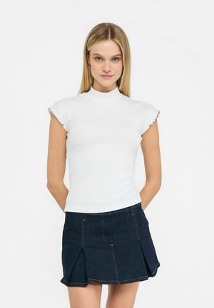 Donna bionda che indossa una maglietta bianca a maniche corte e una mini gonna plisettata in denim scuro, in piedi con le mani dietro la schiena.