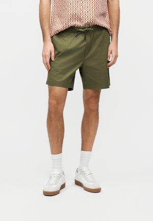 Mężczyzna ubrany w oliwkowozielone szorty ze sznurkiem, białe skarpetki i białe trampki, stojący na tle jednolitego jasnego tła.