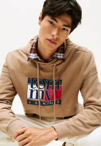 Giovane in felpa beige Tommy Jeans con cappuccio e lacci sopra una camicia a quadri, seduto con le mani unite, sfondo neutro.