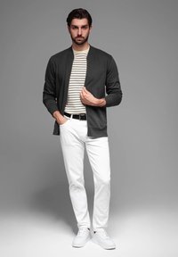 Chaqueta bomber gris oscuro sobre una camisa de rayas blanca y azul marino, combinada con jeans blancos y zapatillas blancas, de pie contra un fondo gris.