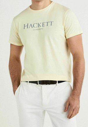 Homme portant un t-shirt Hackett London jaune pâle rentré dans un pantalon blanc avec une ceinture noire, debout avec une main dans la poche.