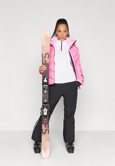 Ροδοκόκκινη μπουφάν, λευκή μπλούζα με μακριά μανίκια, μαύρο παντελόνι σκι. Κρατά καφέ σκι με μαύρες βάσεις. Κοντά μαλλιά σε πλεξούδες. Φόντο στο στούντιο.