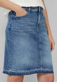 Denim rok in middelblauw met een rafelige zoom, heeft een klassieke vijf-pocket ontwerp en een rechte snit, getoond met een hand in een zak.