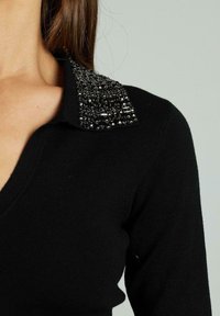 Schwarzer Pullover mit V-Ausschnitt und figurnahem Design. Der Kragen ist mit dekorativen schwarzen Perlen und Pailletten verziert, was Textur und visuelles Interesse hinzufügt.