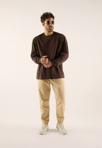 Brun langermet skjorte, beige tilspissede bukser og hvite joggesko; solbriller fullfører det avslappede antrekket. Glatt stoff, avslappet passform.