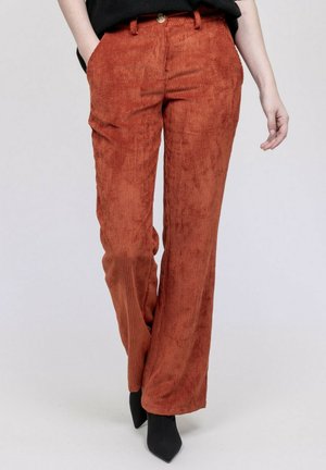 Personne portant un pantalon en velours côtelé couleur rouille avec des poches avant et des bottes noires à bout pointu, debout devant un fond uni.
