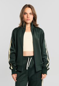 Joven con cabello castaño claro que lleva una chaqueta verde oscuro con cremallera y pantalones a juego con rayas crema, y una camiseta corta crema debajo.