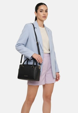 Nő világoskék zakóban, bézs felsőben, levendulaszínű rövidnadrágban, fekete bőr válltáskával sima háttér előtt.