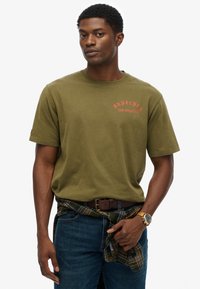 Camiseta de algodón verde oliva con estampado en rojo "Superdry Los Ángeles". Combinada con jeans azul oscuro y un cinturón de cuero marrón. Camisa de franela texturizada atada a la cintura.