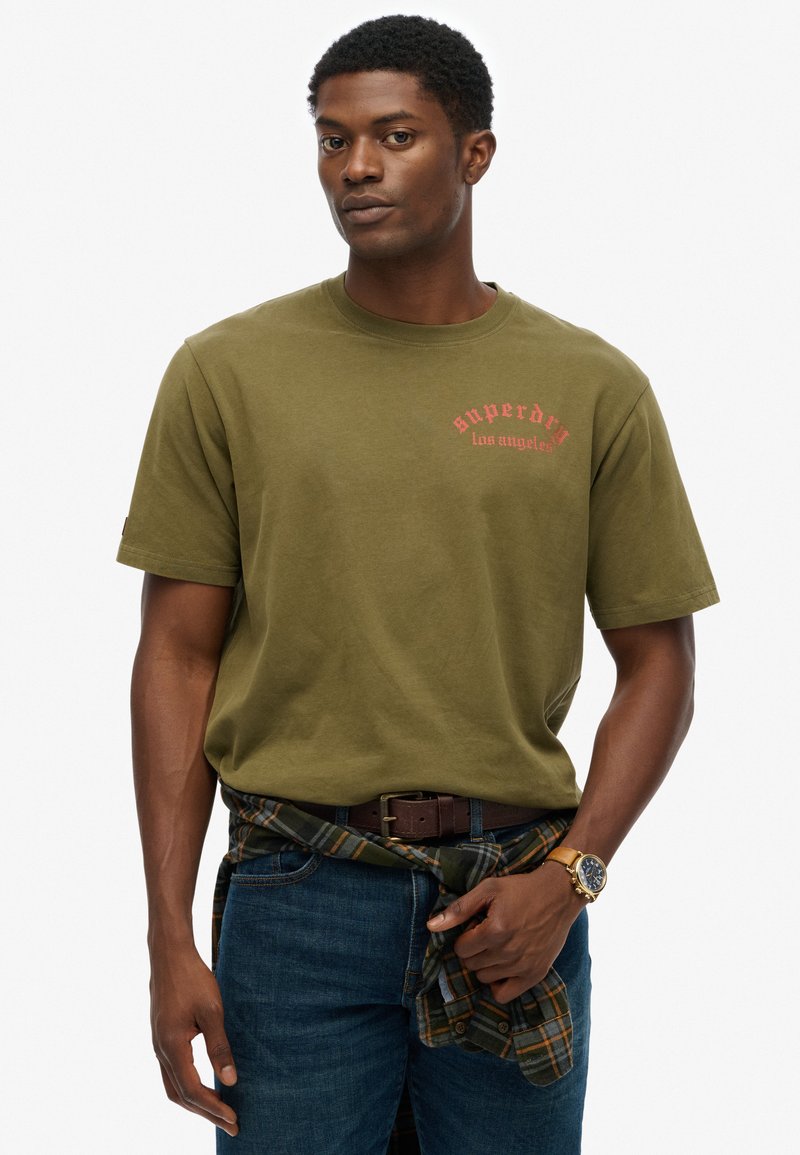 Camiseta de algodón verde oliva con estampado en rojo "Superdry Los Ángeles". Combinada con jeans azul oscuro y un cinturón de cuero marrón. Camisa de franela texturizada atada a la cintura.