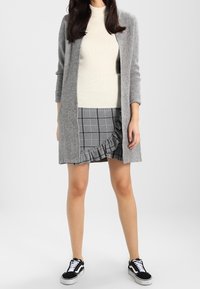 Szary długi kardigan na kremowym, prążkowanym swetrze, w połączeniu z czarno-białą, kratkowaną spódnicą z falbanką na dole oraz czarnymi trampkami.