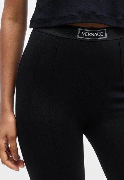 Leggings noirs en tissu extensible, dotés d'une large ceinture élastique avec une étiquette blanche portant le logo "VERSACE". Texture lisse, coupe ajustée.