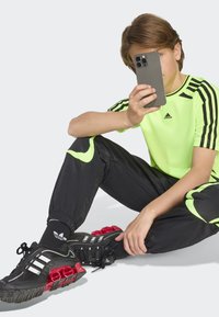 Neon žalia trumpa rankovė marškiniai su juodomis juostelėmis, juodos kelnės su neoniniais akcentais ir juodi sportbačiai su raudonais akcentais. Telefonas laikomas rankoje.