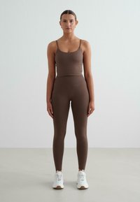 Hnědý sportovní set skládající se z upnutého tílka s tenkými ramínky a legín s vysokým pasem, vyrobený z elastického materiálu, doplněný bílými teniskami.