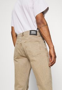 Vaqueros de mezclilla beige con corte recto, que presentan una etiqueta de marca negra en la parte trasera de la cintura y dos bolsillos traseros con una textura sutil.