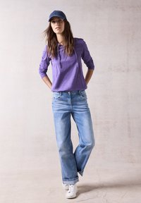 Felpa viola a maniche lunghe con cordino, jeans chiari a vestibilità ampia e sneakers bianche. Outfit casual su uno sfondo neutro.