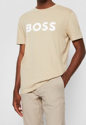 Muž oblečený v béžovém tričku BOSS a světle béžových károvaných kalhotách, s jednou rukou v kapse na neutrálním šedém pozadí.