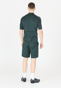 Dunkelgrünes Sportshirt mit kurzen Ärmeln, hohem Kragen und Taschendetails auf der Rückseite, kombiniert mit passenden Shorts. Das Material scheint leicht und atmungsaktiv zu sein.