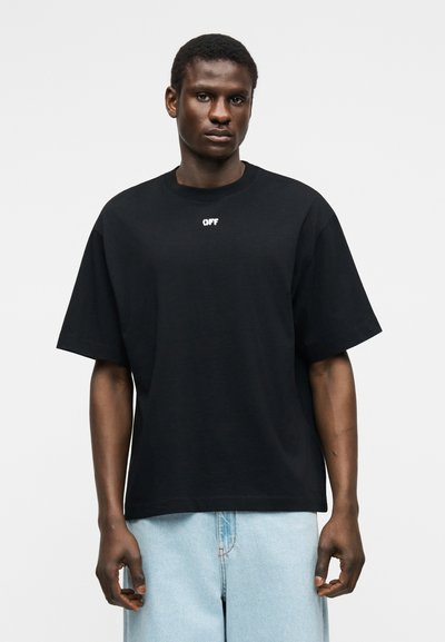 Άνδρας με φαρδιά μαύρη μπλούζα με την επιγραφή «OFF» στο στήθος και ανοιχτόχρωμο τζιν, στέκεται μπροστά σε απλό λευκό φόντο.