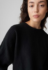 Schwarzer, strukturierter Pullover mit rundem Ausschnitt und weiten Ärmeln. Der Stoff zeigt ein dezentes, erhabenes Muster und wirkt weich.