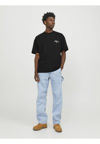 Sort kortærmet t-shirt med hvidt logo på brystet, parret med lyseblå jeans og brune støvler, stående mod en ensfarvet baggrund.