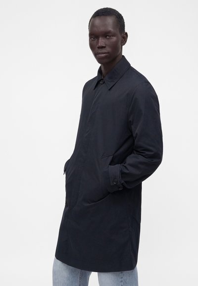 Jeune homme portant un long manteau bleu marine foncé, les mains dans les poches, debout devant un fond blanc uni.