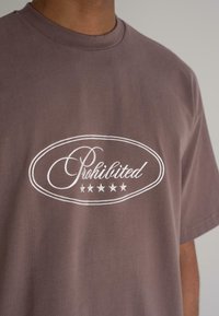 Short-sleeve brown cotton t-shirt featuring a white cursive "Prohibited" logo within an oval frame, accented by five stars below.