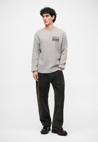 Szary longsleeve z teksturowanym wzorem i czarnym logo na klatce piersiowej, w połączeniu z ciemnymi, luźnymi spodniami i czarnymi butami.