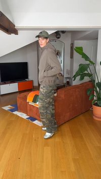 Νεαρός άνδρας με πράσινο καπέλο, μπεζ μπουφάν, παραλλαγής παντελόνι cargo και ασημένια παπούτσια στέκεται σε ένα σύγχρονο σαλόνι με ξύλινο δάπεδο και πορτοκαλί καναπέ.