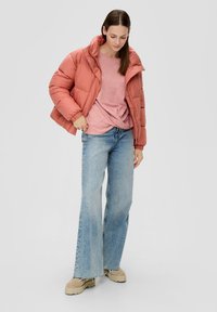 Ein rostfarbener Pufferjacke, ein hellrosa Oberteil, weit ausgestellte blaue Jeans und beigefarbene Plateauschuhe. Das Outfit hat einen lässigen, lockeren Schnitt.