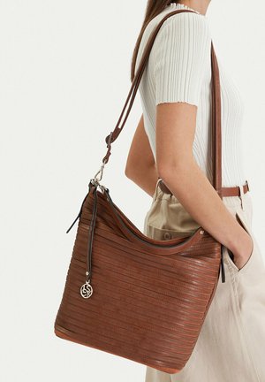 Žena v bílém žebrovaném tričku s krátkým rukávem a béžových kalhotách drží velkou hnědou koženou kabelku s texturovaným proužkovým vzorem.