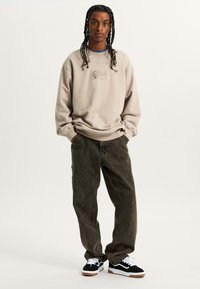 Beige sweatshirt with embroidered logo on chest, paired with dark green jeans and black sneakers featuring white laces and brown soles.